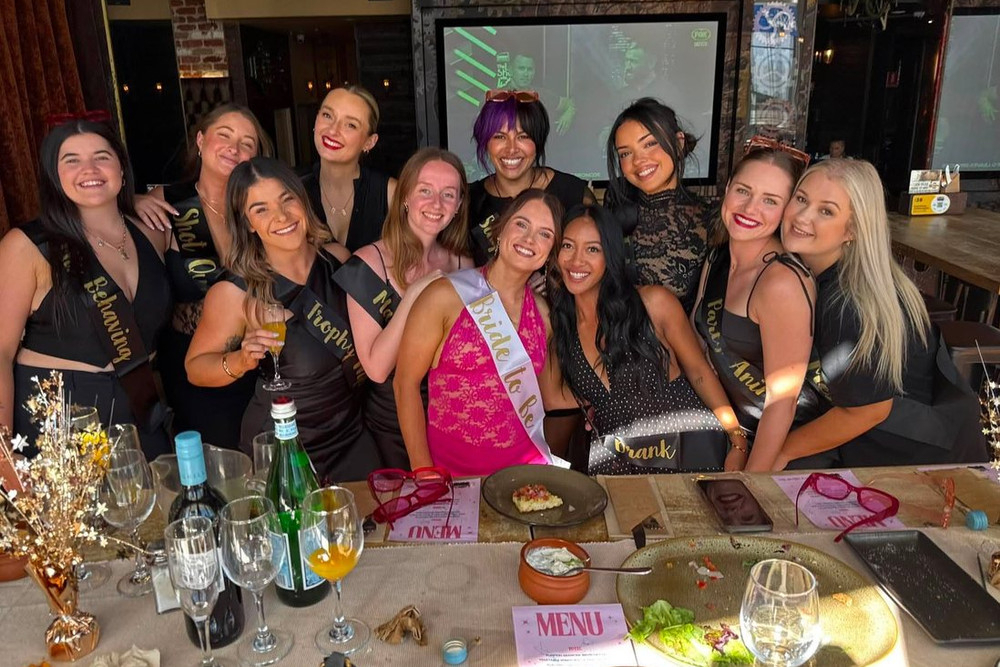 A big hens group in sashes gathered behind a long lunch table of plates and drinks