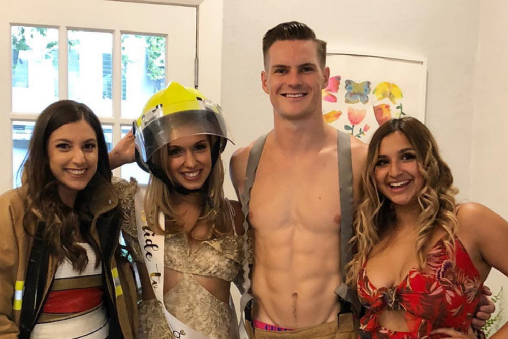 The bride wearing a fireman's helmet beside a shirtless fireman, her friends grinning either side