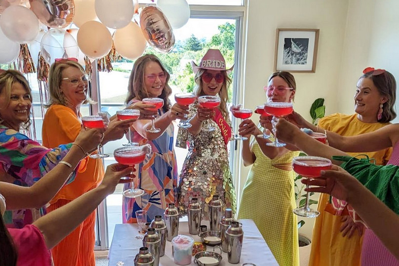 Hens in bright dresses raising pink cocktails over a long table of shakers