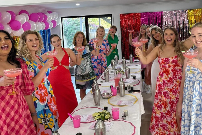 A hens crew in summer dresses raising pink cocktails around a long table