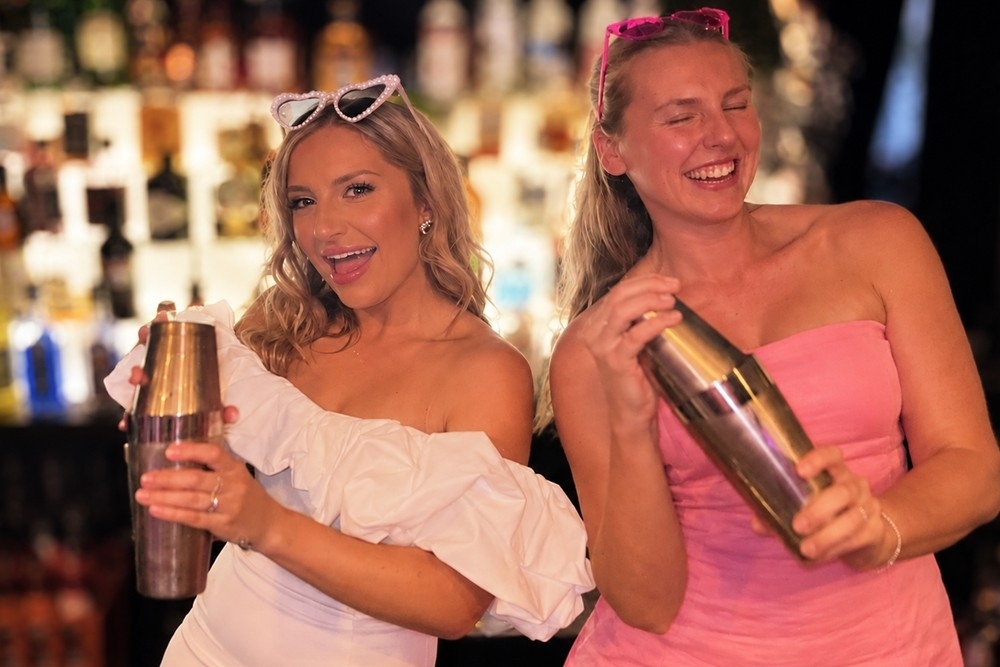 Two hens laughing as they shake cocktails in front of a glowing back bar