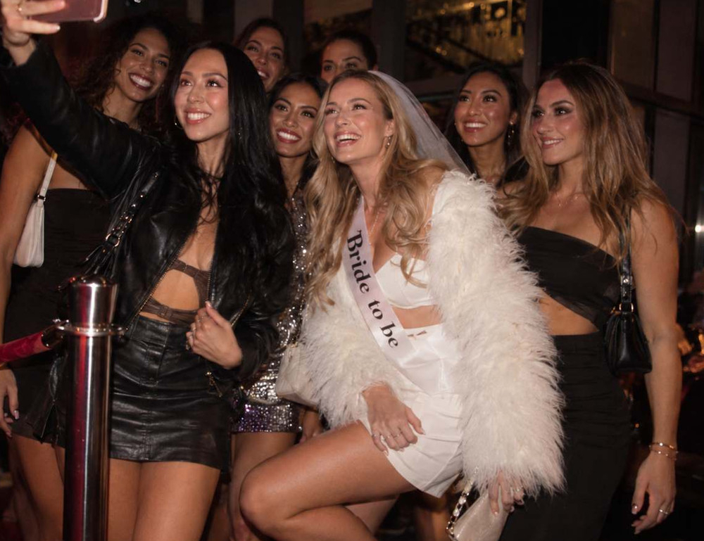 The bride in a sash and feather jacket laughing in a selfie with her crew on a night out