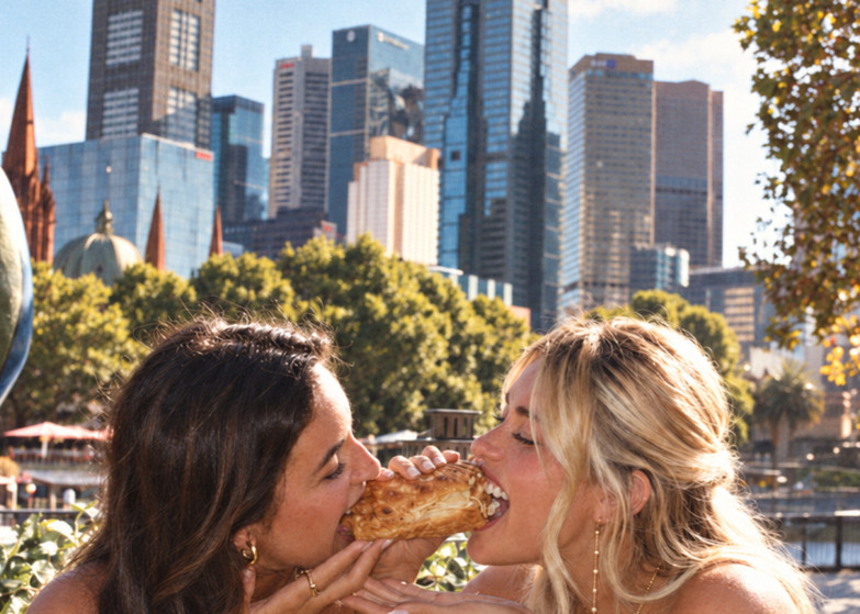 Two friends laughing as they share one pastry, the Melbourne skyline behind them
