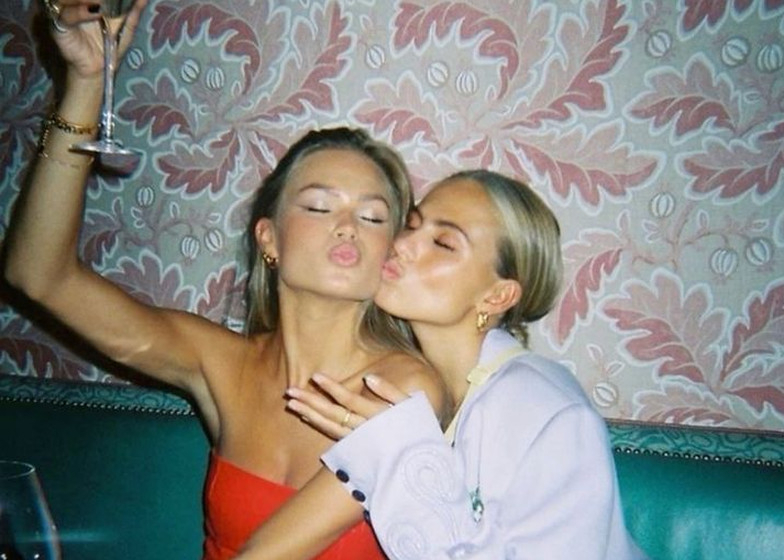 Two friends blowing kisses with a glass of bubbly raised in a velvet booth