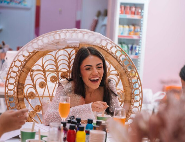 A laughing bride to be in a peacock chair with a spritz, paint bottles lined up on the class table