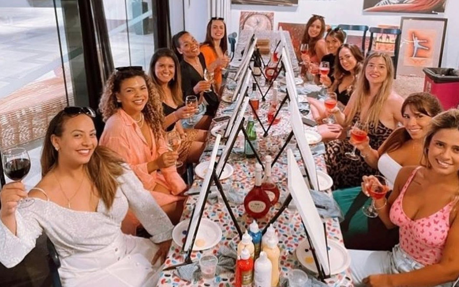 Friends raising their wine glasses along a long table of easels and canvases