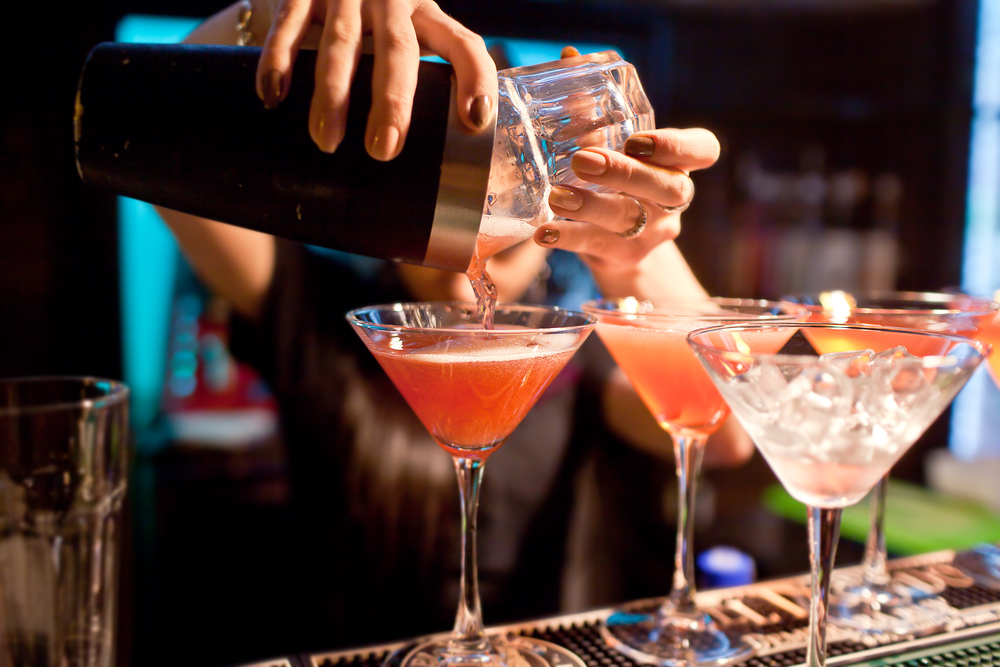 A bartender pours pink cocktails from a shaker into a row of martini glasses