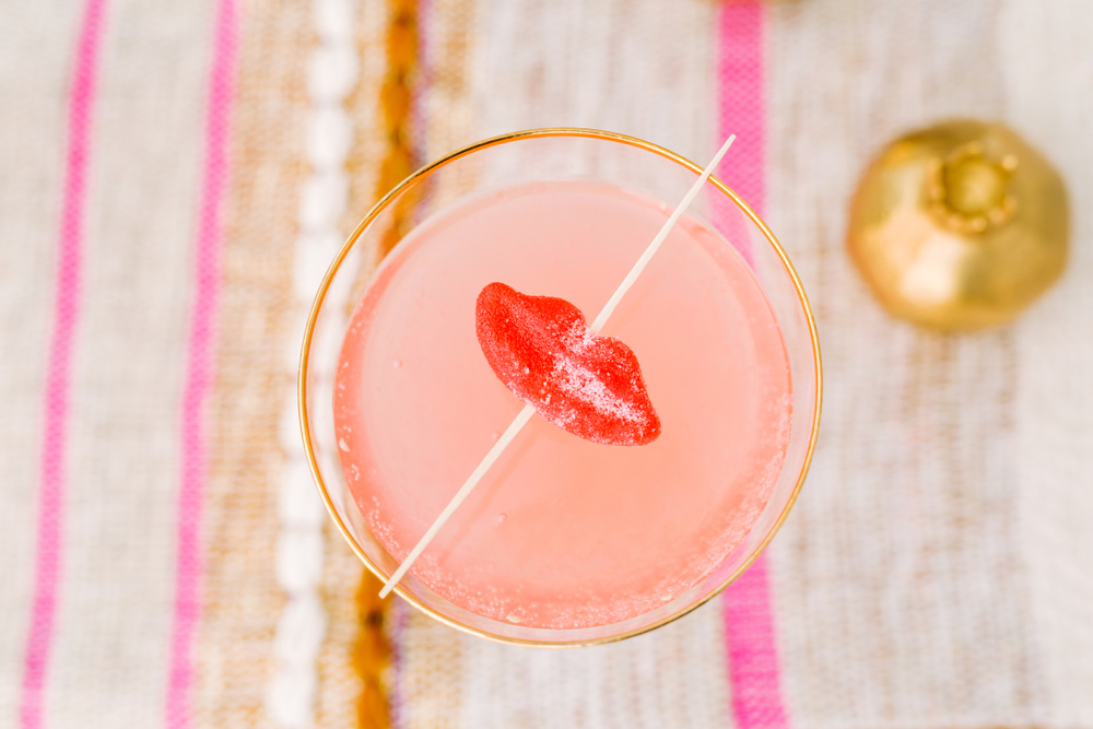 A pink cocktail in a coupe with a red lips garnish on a striped cloth
