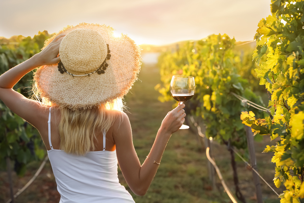 A woman in a straw hat holding up a glass of wine in a sunlit vineyard