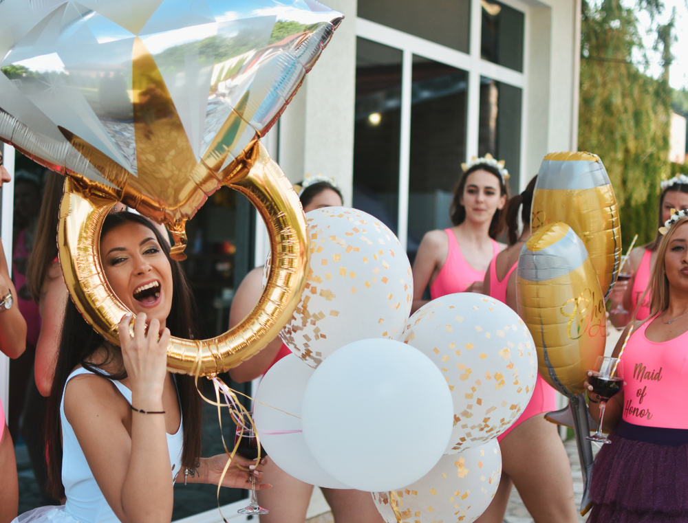 A laughing woman holding a giant ring balloon with friends and white balloons around her