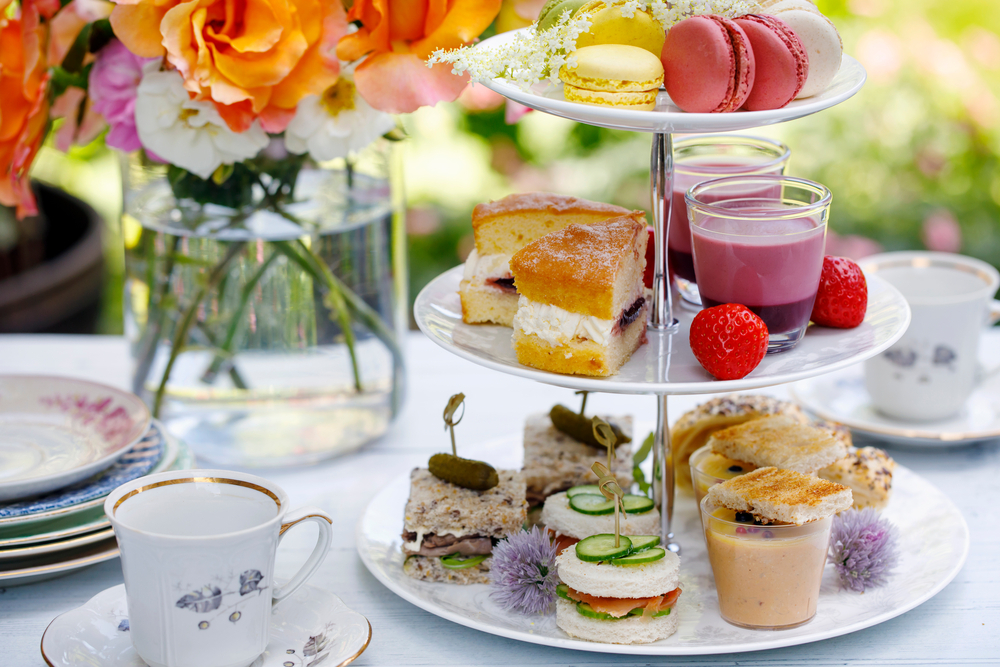 A tiered high tea stand with macarons, sandwiches and cakes beside teacups and flowers